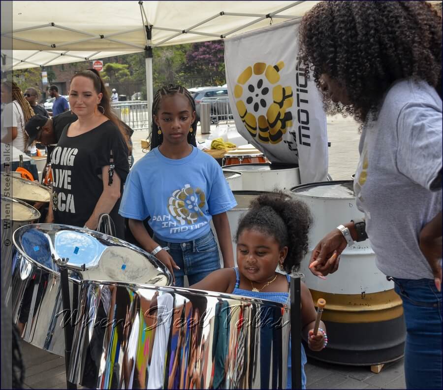 The youngest to step up to experience ‘playing pan’ during the second workshop—this time with Pan In Motion—as musicians look on