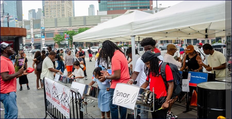 Steelpan musician (left) keeps time/rhythm on cow bell - while the ‘Newbies’ try their hands at performing a very short tune after a quick hands-on workshop, with Eruption Steel Orchestra players looking over them during the Counterpoint: Brooklyn Steel Pan Fête